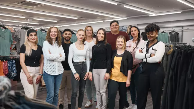 Un gruppo di collaboratori posa insieme in un punto vendita KiK, davanti agli espositori di abbigliamento, guardando verso la fotocamera.