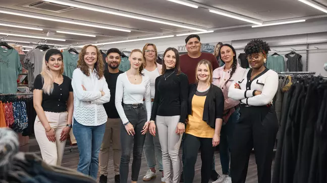 Un gruppo di collaboratori si trova in un punto vendita KiK davanti agli espositori di abbigliamento e guarda verso la fotocamera.