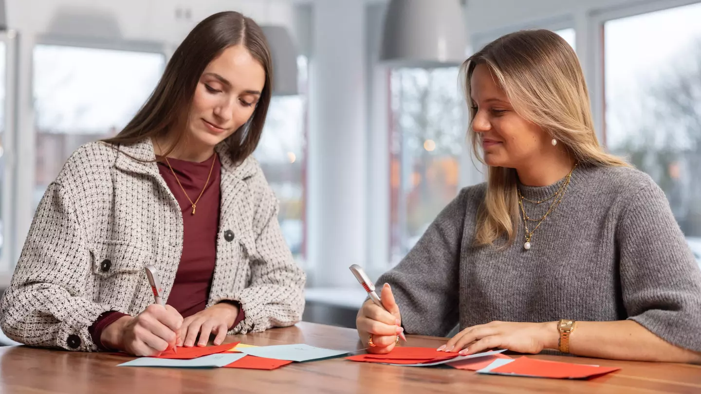 Zwei Mitarbeiterinnen stehen an einem Tisch mit Kugelschreiber in der Hand und schauen auf die Unterlagen die auf dem Tisch liegen