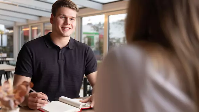 Ein Mann und eine Frau schauen in ein Notizbuch, die Frau hält einen Kugelschreiber, beide lächeln und sitzen in einem modernen Büro