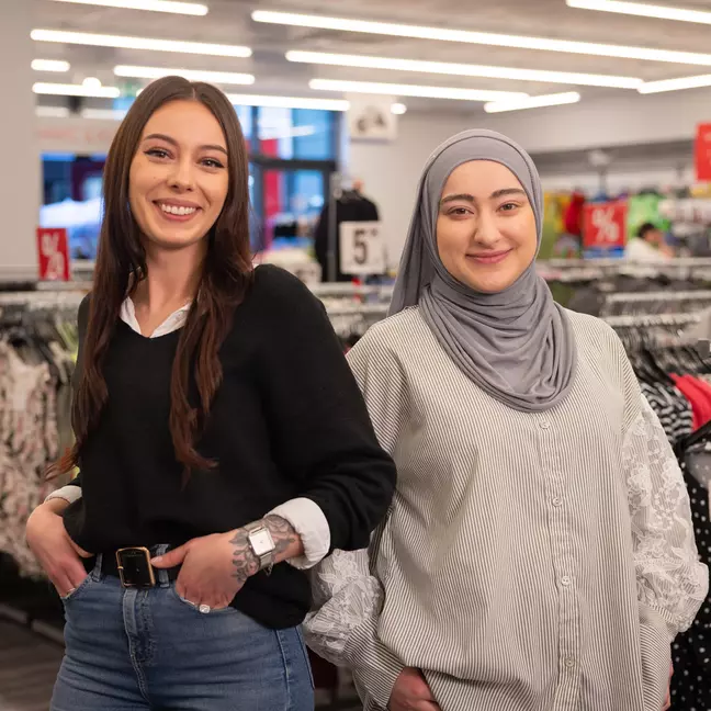 Due dipendenti sorridono davanti a degli espositori di abbigliamento in un negozio KiK