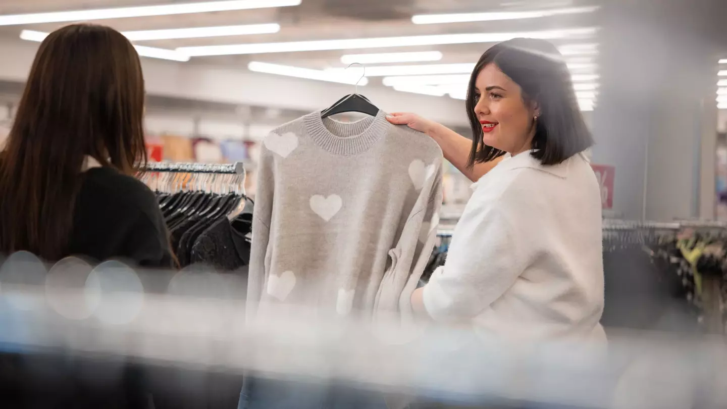 Una collaboratrice mostra a una cliente un maglione grigio con motivo a cuore all’interno di un negozio di abbigliamento.