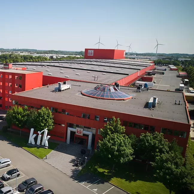 Un grande edificio aziendale rosso con il logo KiK si erge in un paesaggio verdeggiante.
