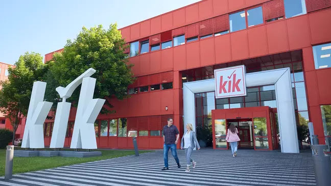 Un edificio aziendale rosso con un grande logo bianco KiK all’ingresso, mentre diverse persone si dirigono verso l’edificio.